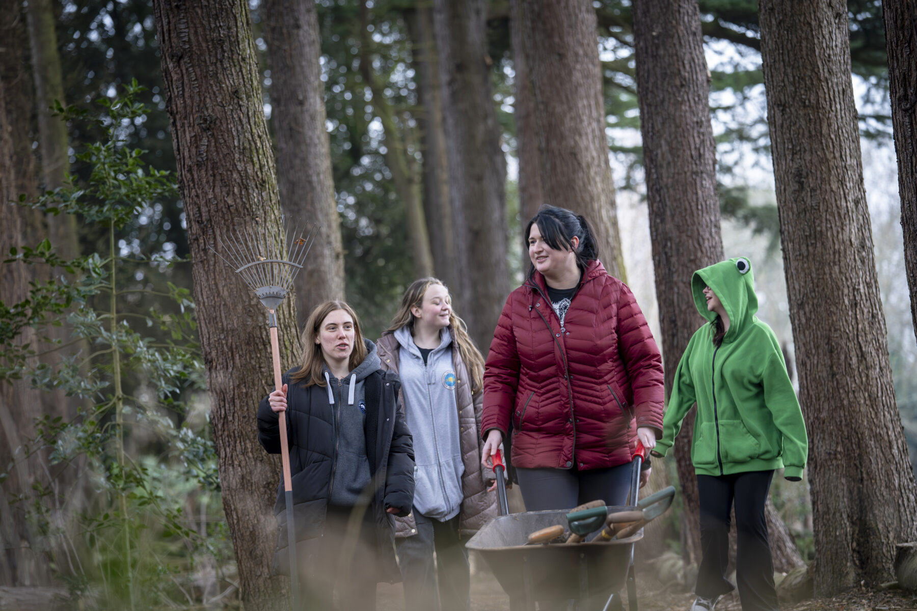 A group of women students walk through a wood, one is pushing a wheelbarrow